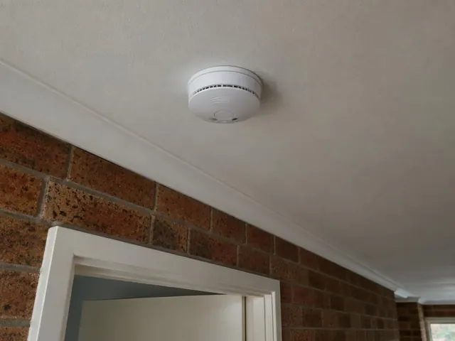 Ceiling-mounted smoke alarm above a brick hallway wall and doorway in Belmont